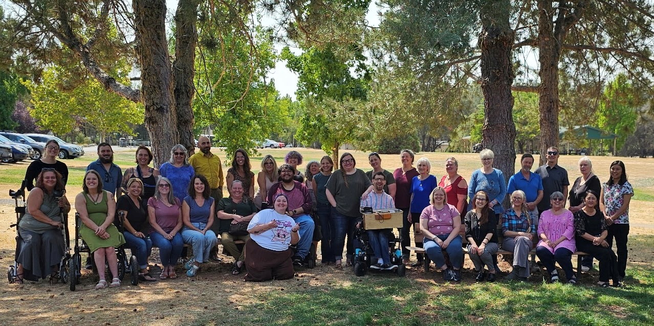 FREED staff gathered under tall trees on a sunny day. Some seated, some standing, some use mobility aids.